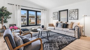 Ein modernes Wohnzimmer mit einem beigefarbenen Sofa, einem runden Couchtisch und einem gemusterten Teppich. Ein großes Fenster mit Vorhängen, eine Topfpflanze und Kunstwerke an der Wand sind ebenfalls zu sehen.