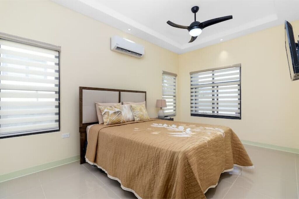 A bedroom with a bed, ceiling fan, air conditioning unit, and windows with blinds.