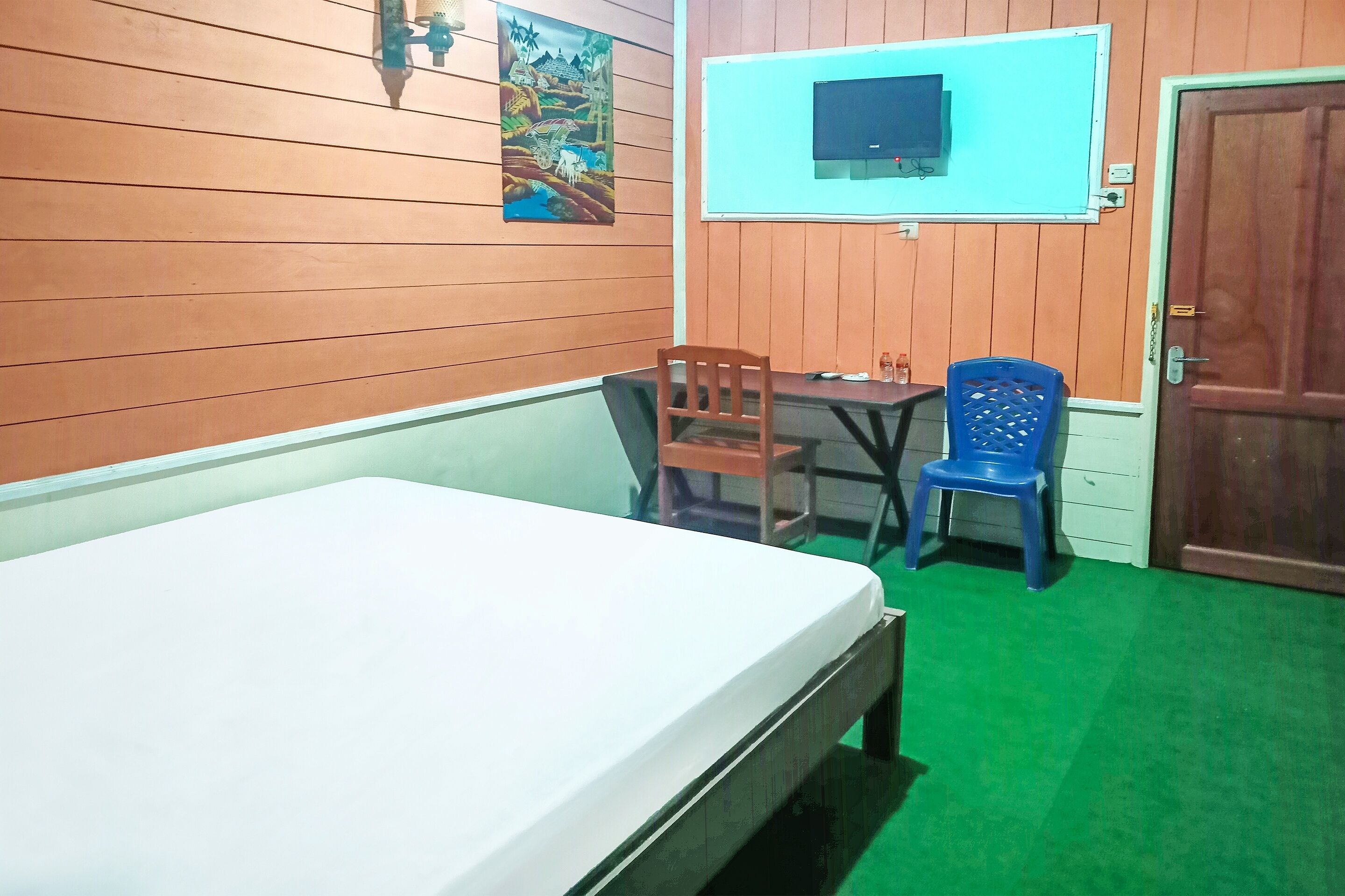 A room with a bed, a table, a blue chair, a TV mounted on the wall, and a door.