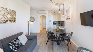 Ein modernes Wohnzimmer mit einem grauen Sofa, einem runden Esstisch mit Drahtstühlen und einem an der Wand montierten Flachbildfernseher.