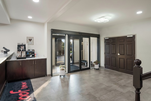 A hotel lobby with a reception desk, a coffee station, a sliding glass door leading to a room, and a dark wooden door. - Red Roof Inn & Suites Carrollton, Ga-west Georgia (Carrollton)