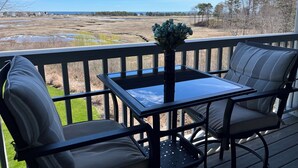A deck with a table and two chairs, overlooking a coastal landscape with fields and trees.