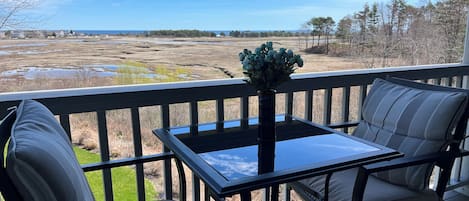 A deck with a table and two chairs, overlooking a coastal landscape with fields and trees.