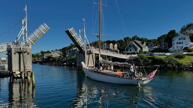 A sailboat docked at a pier with a bridge overhead, surrounded by houses and calm water.