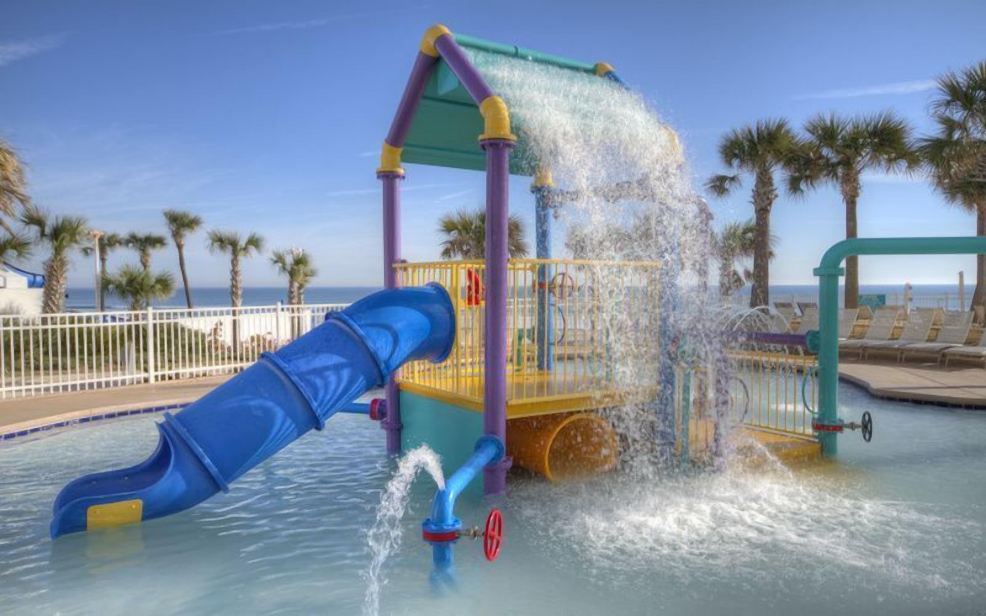 Zona de juegos acuáticos con un tobogán azul y chorros de agua, rodeada de palmeras y un área de descanso junto a la piscina.