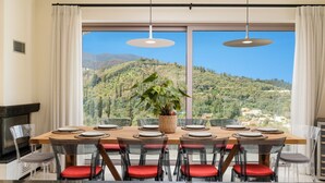Un espace repas avec une grande table, des chaises rembourrées de couleur rouge et une vue sur un paysage montagneux grâce à de grandes fenêtres.