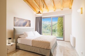 Une chambre avec un grand lit, une table de chevet, un tableau, une fenêtre donnant sur des arbres et des collines, et un plafond en bois.