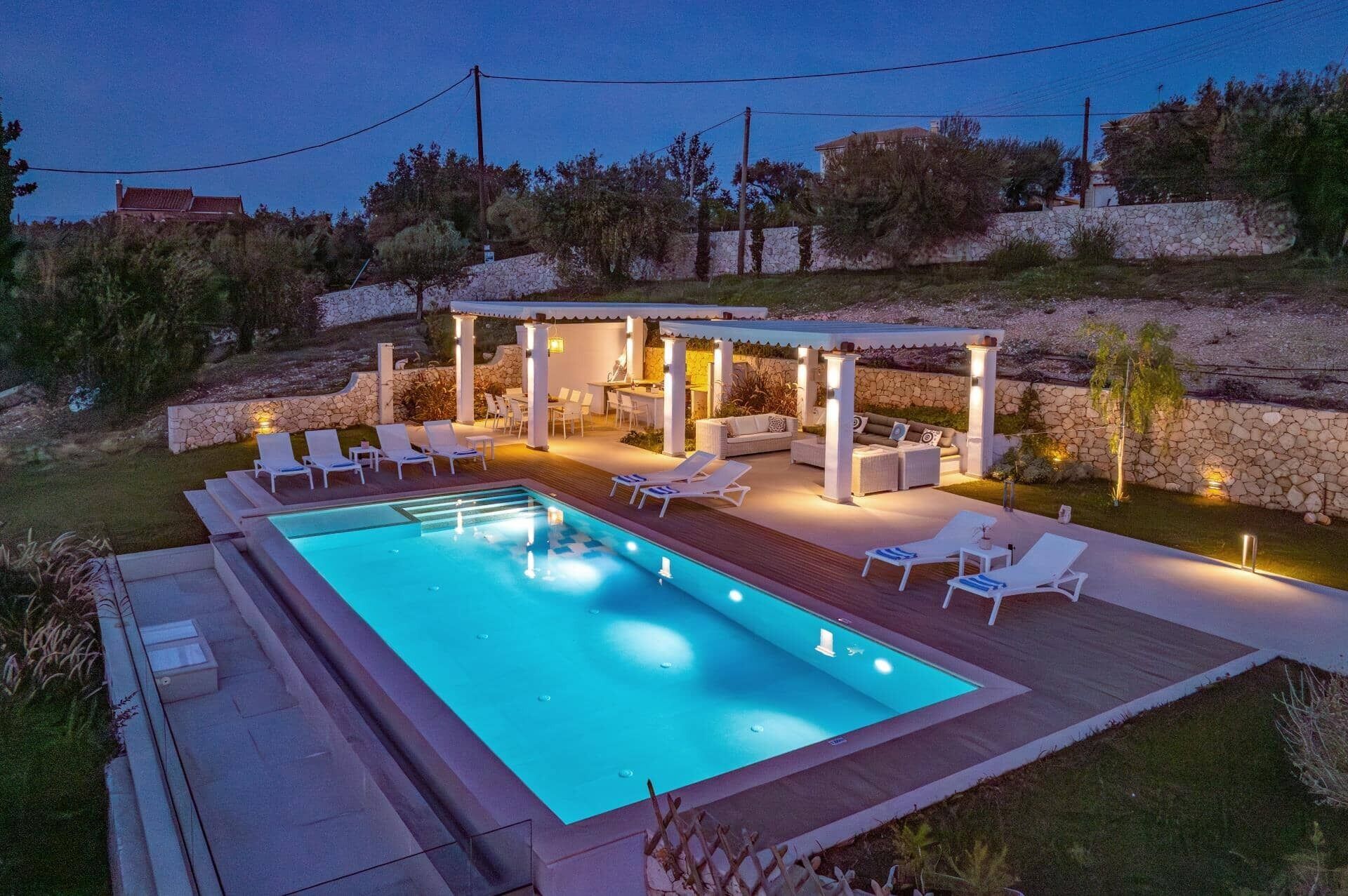 Une piscine rectangulaire dotée de chaises longues et d’un espace de détente couvert, agrémenté de piliers et d’éclairages.