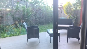 Un patio al aire libre con mobiliario de mimbre, una mesa de vidrio y vistas a un jardín con diversas plantas y una pared.
