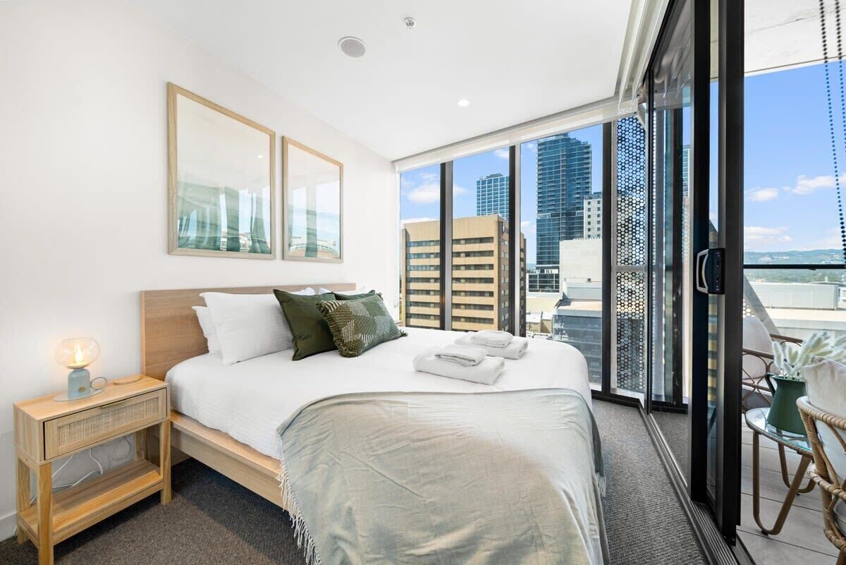 A modern hotel room with a large bed, a nightstand, a window with a city view, and a balcony with a chair and potted plant.
