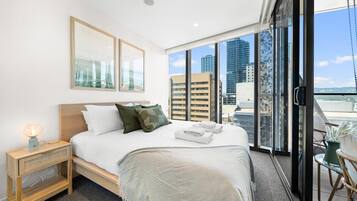 A modern hotel room with a large bed, a nightstand, a window with a city view, and a balcony with a chair and potted plant.