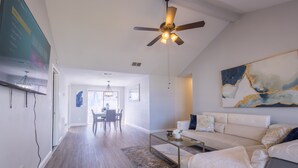 A spacious living room with a white sofa, a glass coffee table, and a ceiling fan. A dining area is visible in the background with a chandelier. - Sleeps 6 Hot Tub Fenced Yard Cozy Chic (Tampa)