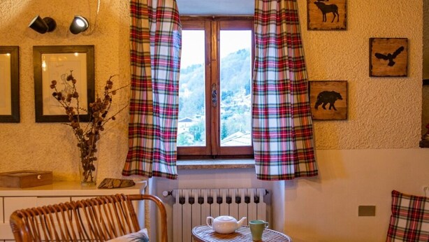 A cozy room with a wooden ceiling, a window with plaid curtains, a wicker chair with a white cushion, a small round table with a teapot and cup, and a radiator. - The mountain loft with the most beautiful park on Lake Maggiore as its garden (TRAREGO VIGGIONA)