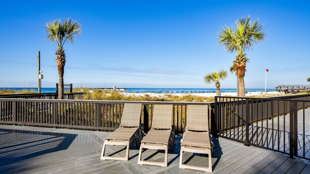 A wooden deck with three wicker lounge chairs facing a beach.