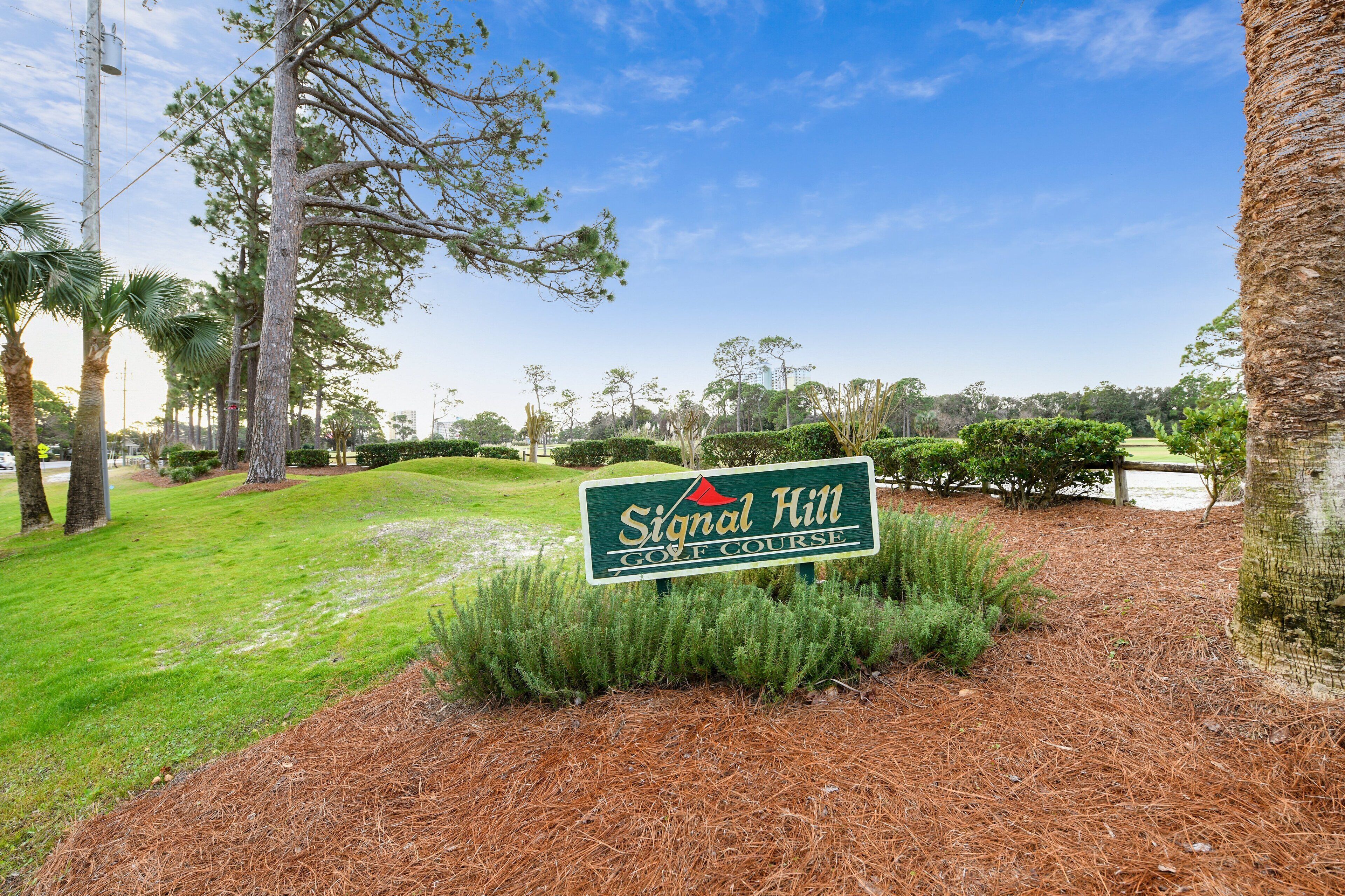A golf course sign with a red triangle logo, surrounded by pine trees and a clear sky.
