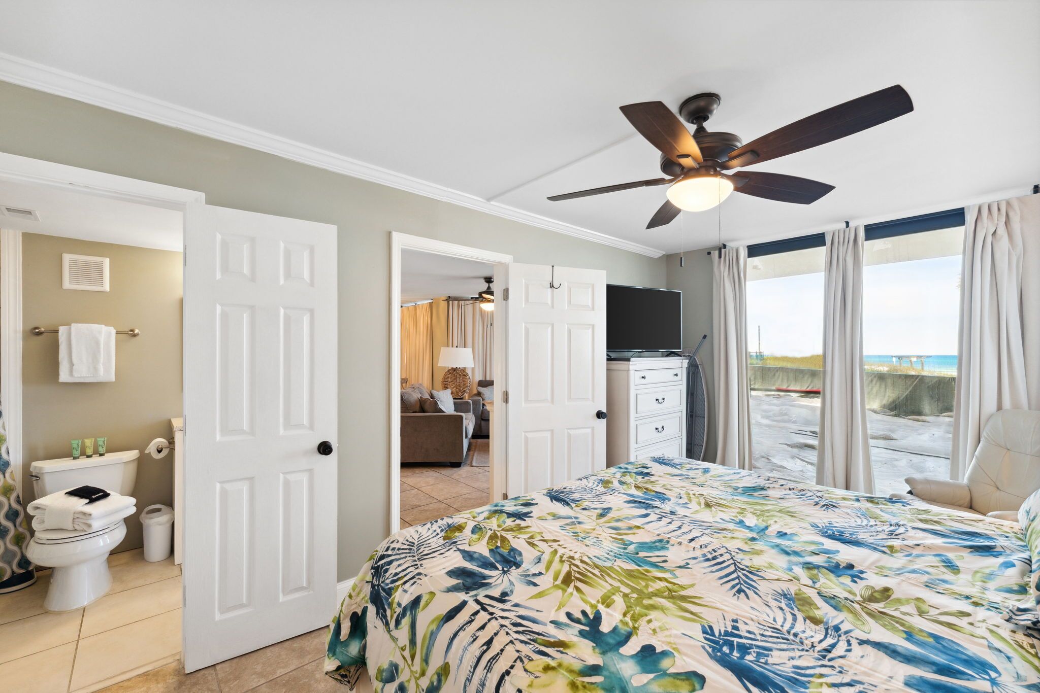 Un dormitorio con cama, ventilador de techo, baño con inodoro y lavabo, y vista a la playa desde la ventana.