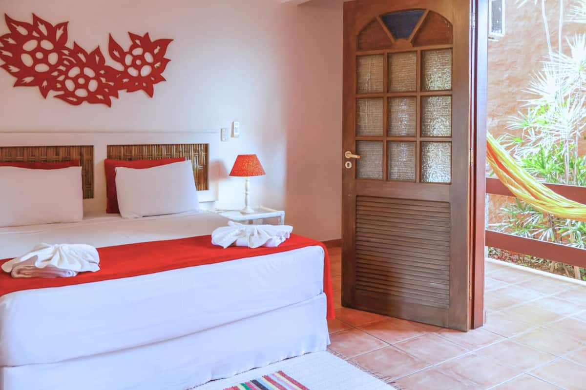Habitación de hotel con cama, mesita de noche, lámpara roja, puerta de madera y vista a un jardín.