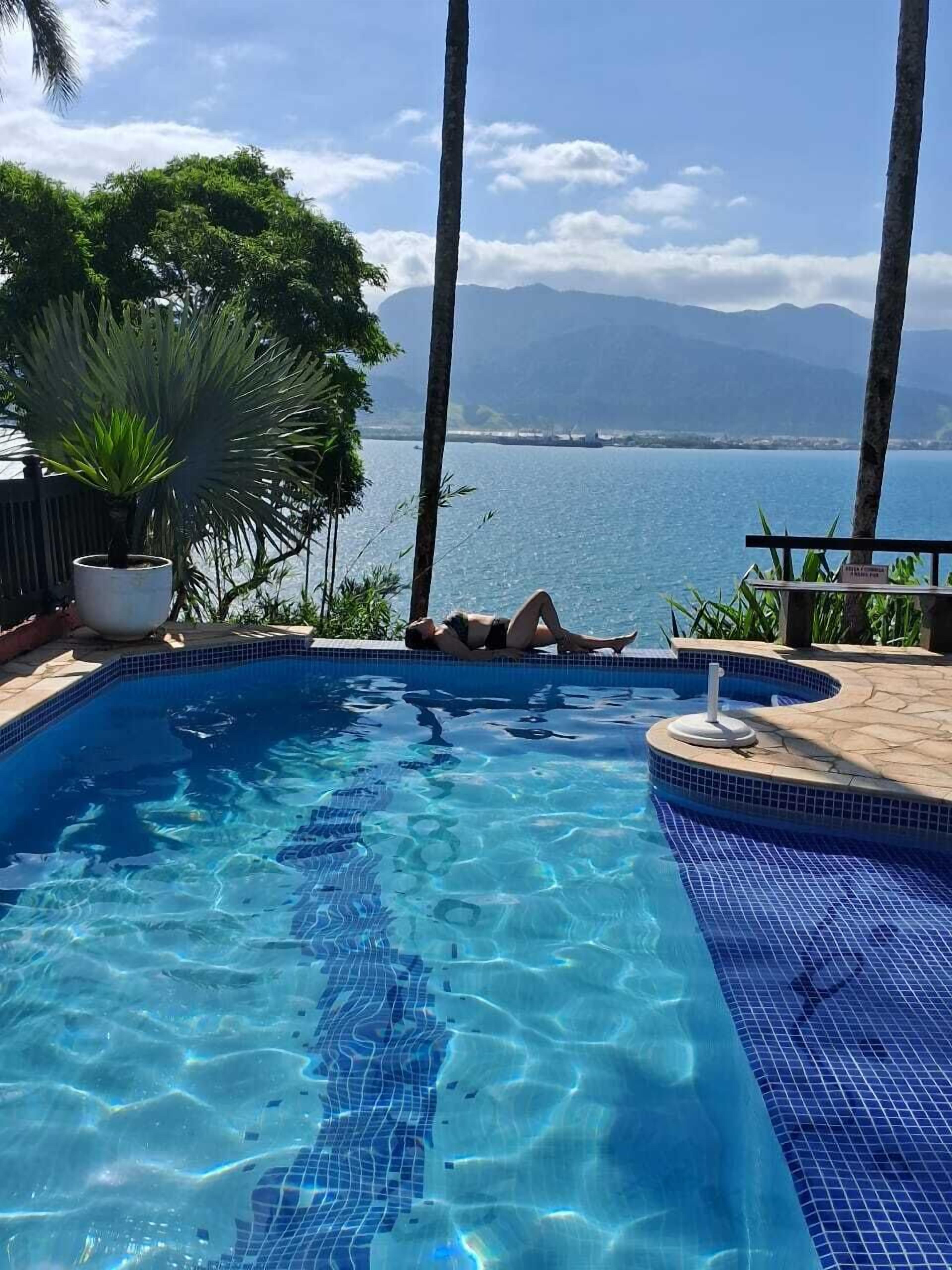 A clear blue swimming pool with tiled edges, surrounded by palm trees and a view of the sea and mountains.
