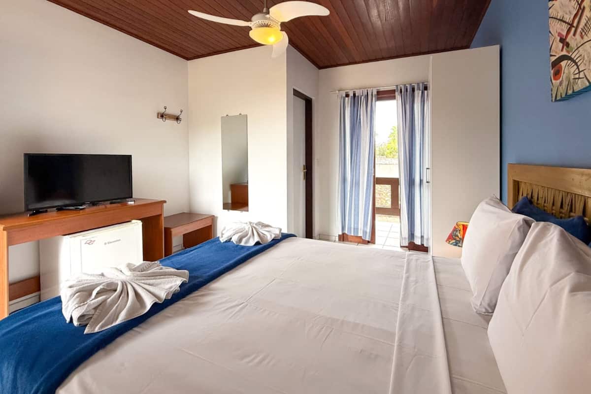 Habitación de hotel con cama, televisor, ventilador de techo, ventana con cortinas y una obra de arte en la pared.