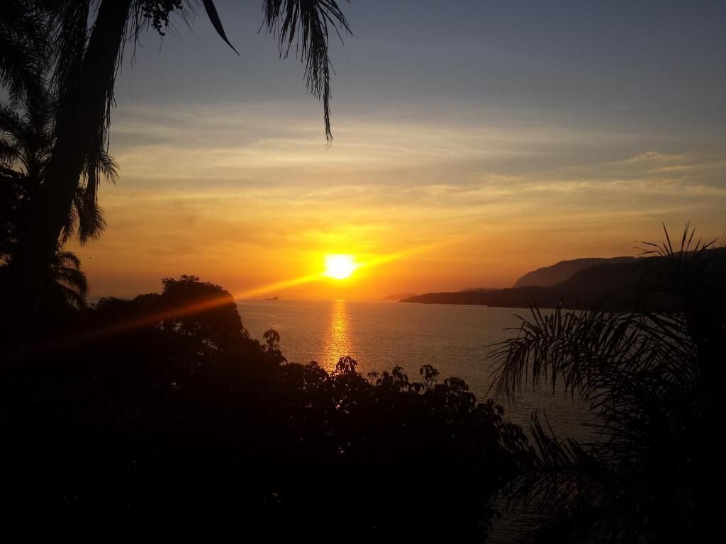 Un atardecer sobre un cuerpo de agua con siluetas de palmeras y colinas distantes.