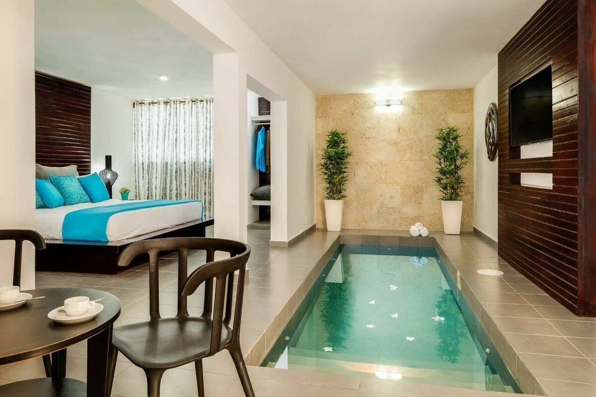 Une chambre d’hôtel moderne avec un lit, une petite table et des chaises, une piscine et deux plantes en pot.