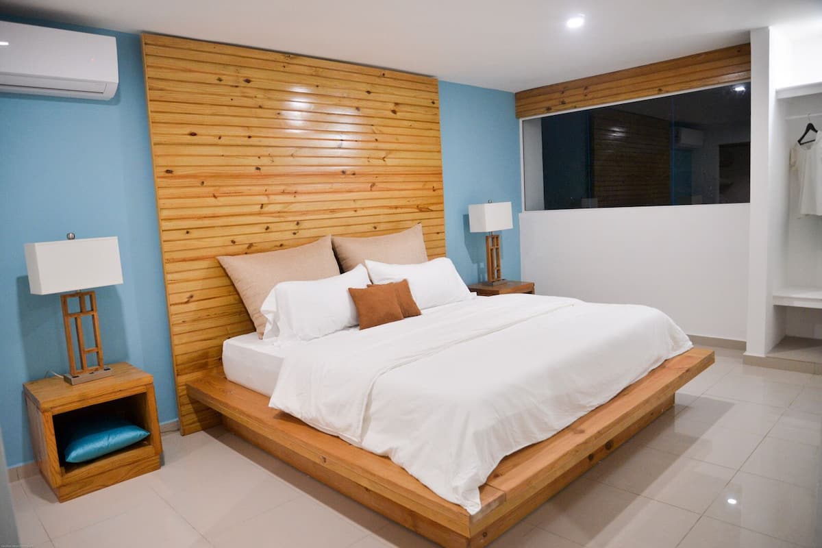 Une chambre moderne avec une tête de lit en bois, un grand lit, deux tables de chevet avec des lampes et une vue sur un ciel sombre par la fenêtre.
