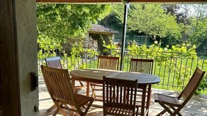 Een overdekt terras met een stenen vloer, houten tafel en stoelen, en uitzicht op groen en een stenen gebouw op de achtergrond.