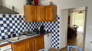 Een keuken met houten kastjes, een spoelbak, een fornuis en een vaatwasser. Er is een eethoek met een tafel en stoelen.
