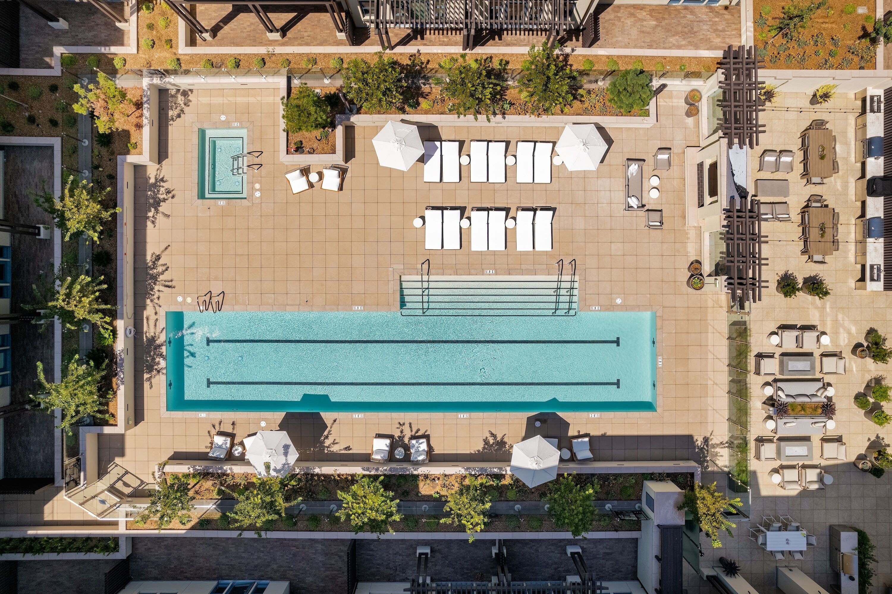 An aerial view of a rooftop pool area with multiple sun loungers, a central swimming pool, and surrounding greenery.