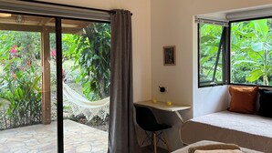 A hotel room with a large bed, a desk with a chair, a ceiling fan, and a view of lush greenery through the windows.