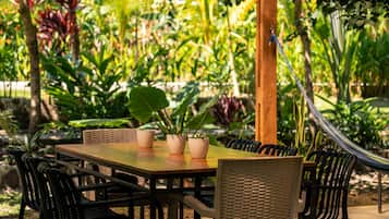 An outdoor dining area with a wooden table and chairs, surrounded by lush greenery and a hammock.