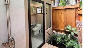 A bathroom with a shower, toilet, and a glass door leading to a wooden deck area with plants.