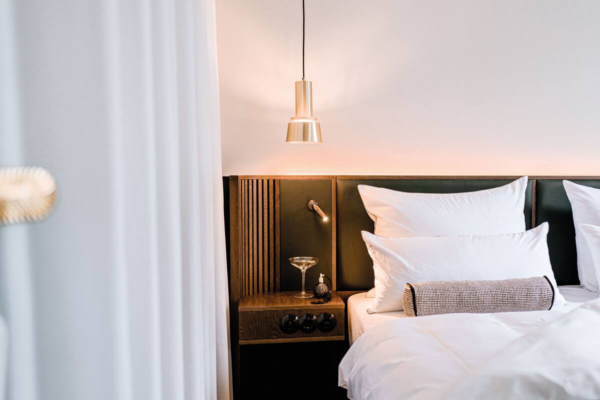 A neatly made bed with white linens, a wooden nightstand with a lamp, and a hanging light fixture.
