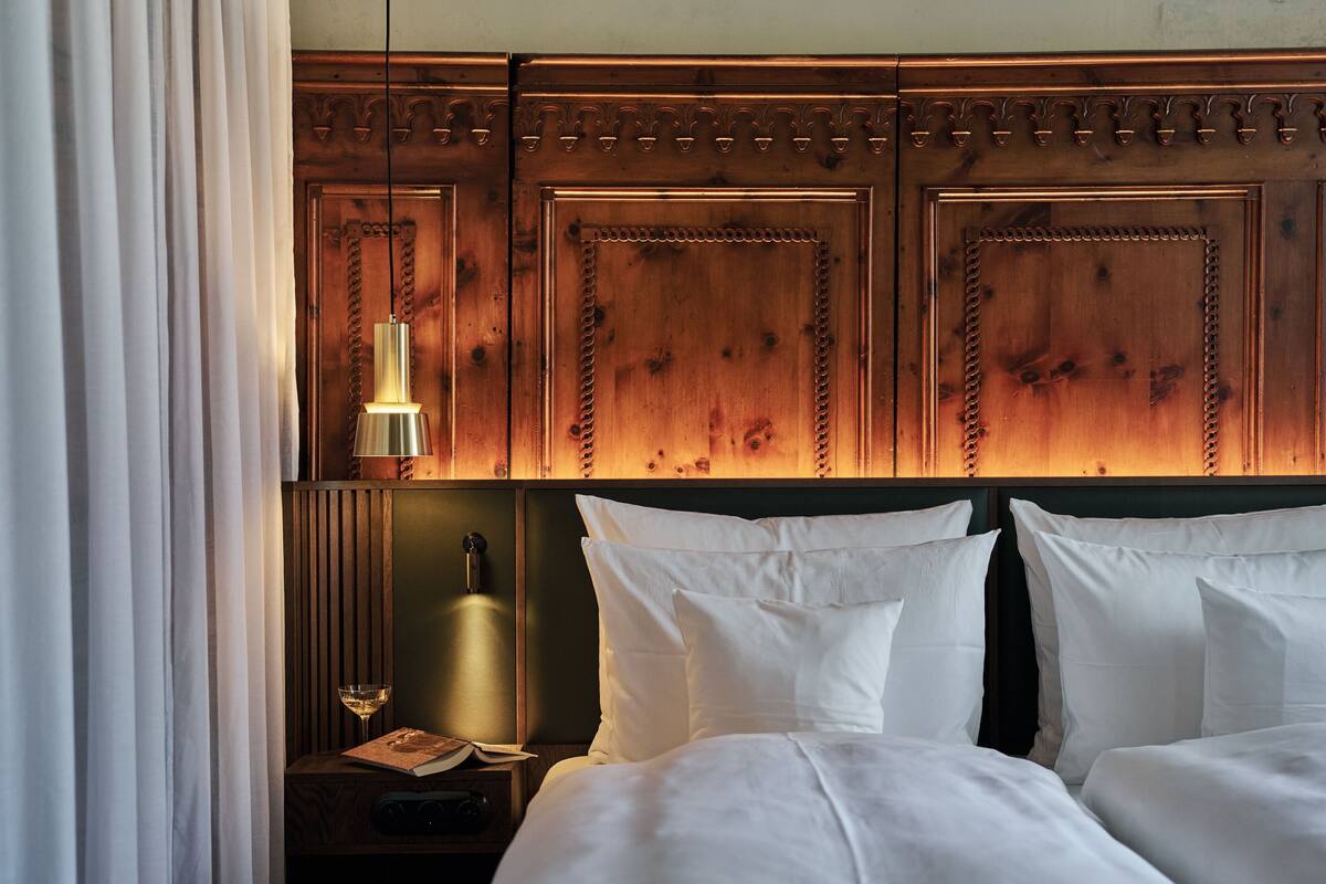 A hotel room with a wooden headboard, two beds, a nightstand with a lamp, a book, and a glass of wine.