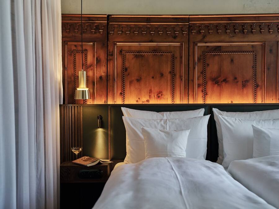 A hotel room with a wooden headboard, two beds, a nightstand with a lamp, a book, and a glass of wine.