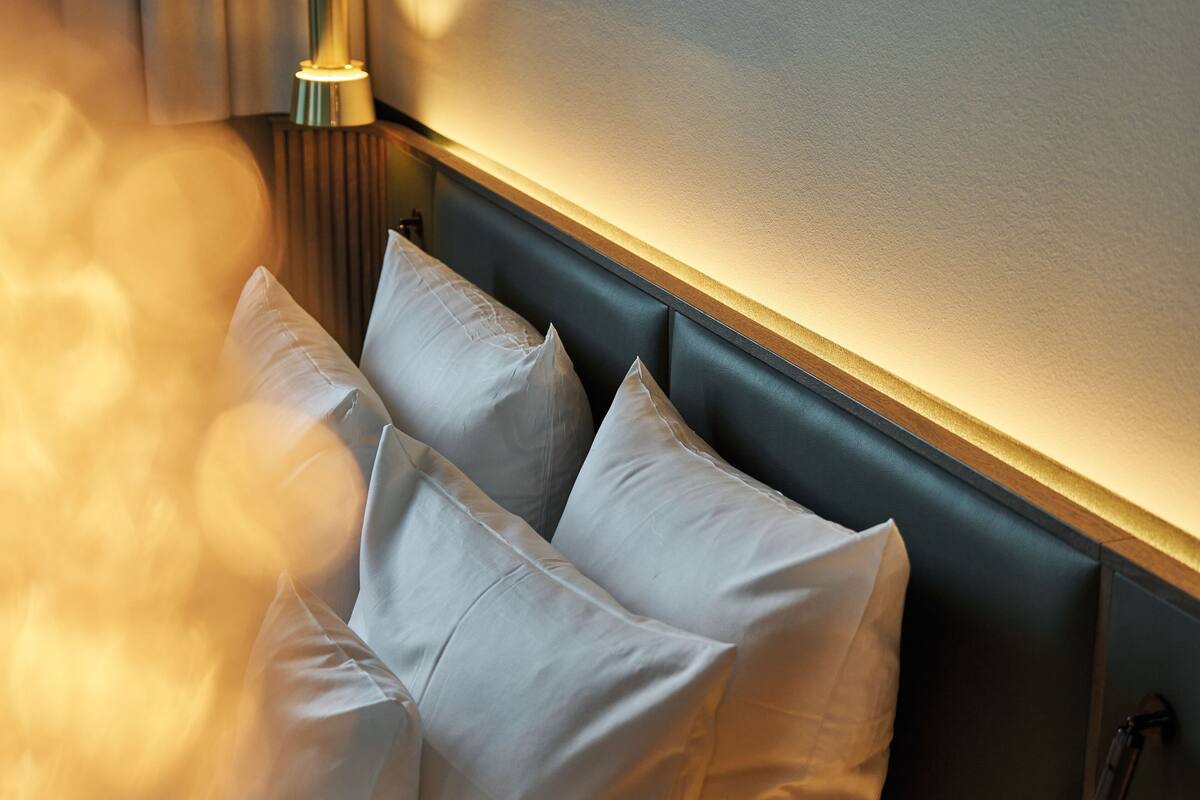 A hotel room with a dark headboard, white pillows, and a wall-mounted light fixture.