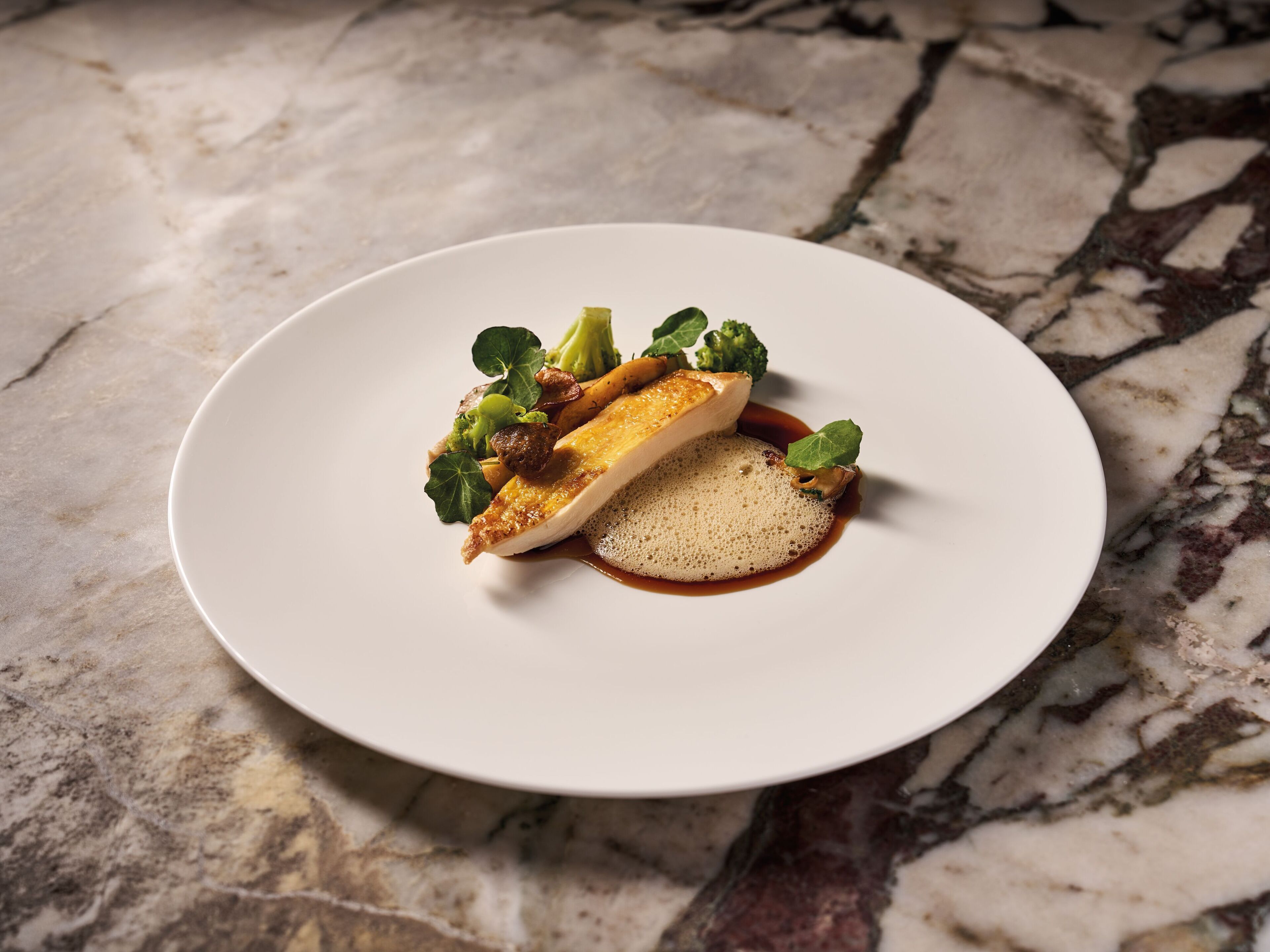 A white plate with a piece of fish, vegetables, and a sauce on a marble surface.