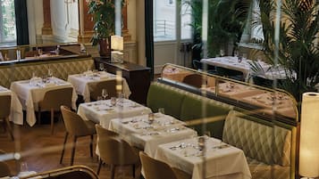 A restaurant with multiple tables set for dining, a green cushioned bench, and large windows allowing natural light.
