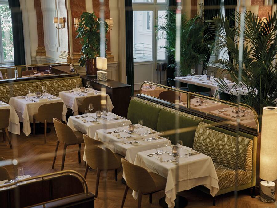 A restaurant with multiple tables set for dining, a green cushioned bench, and large windows allowing natural light.