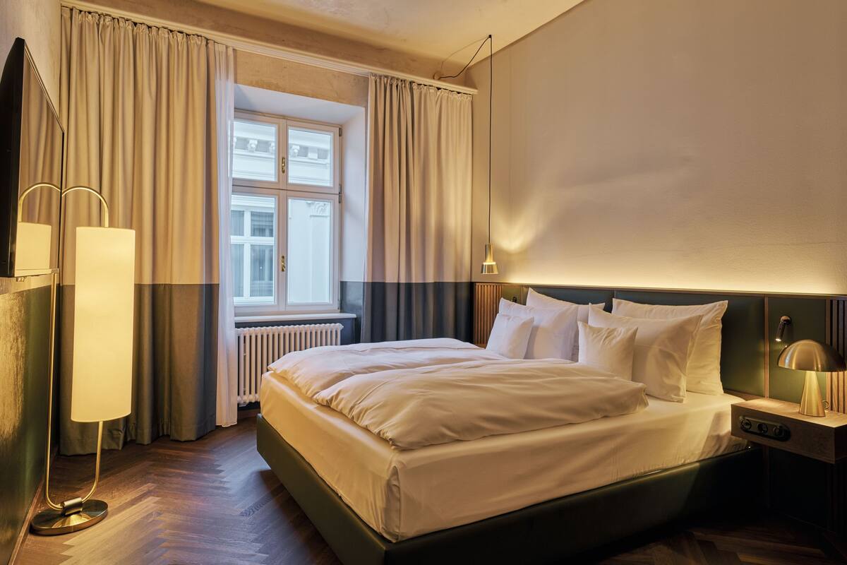 A hotel room with a large bed, a bedside table with a lamp, a window with curtains, and a radiator.