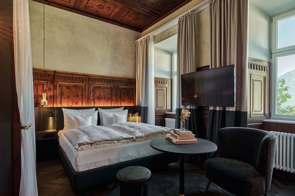 A hotel room with a large bed, a small round table, a chair, a TV, and a view of the outdoors.