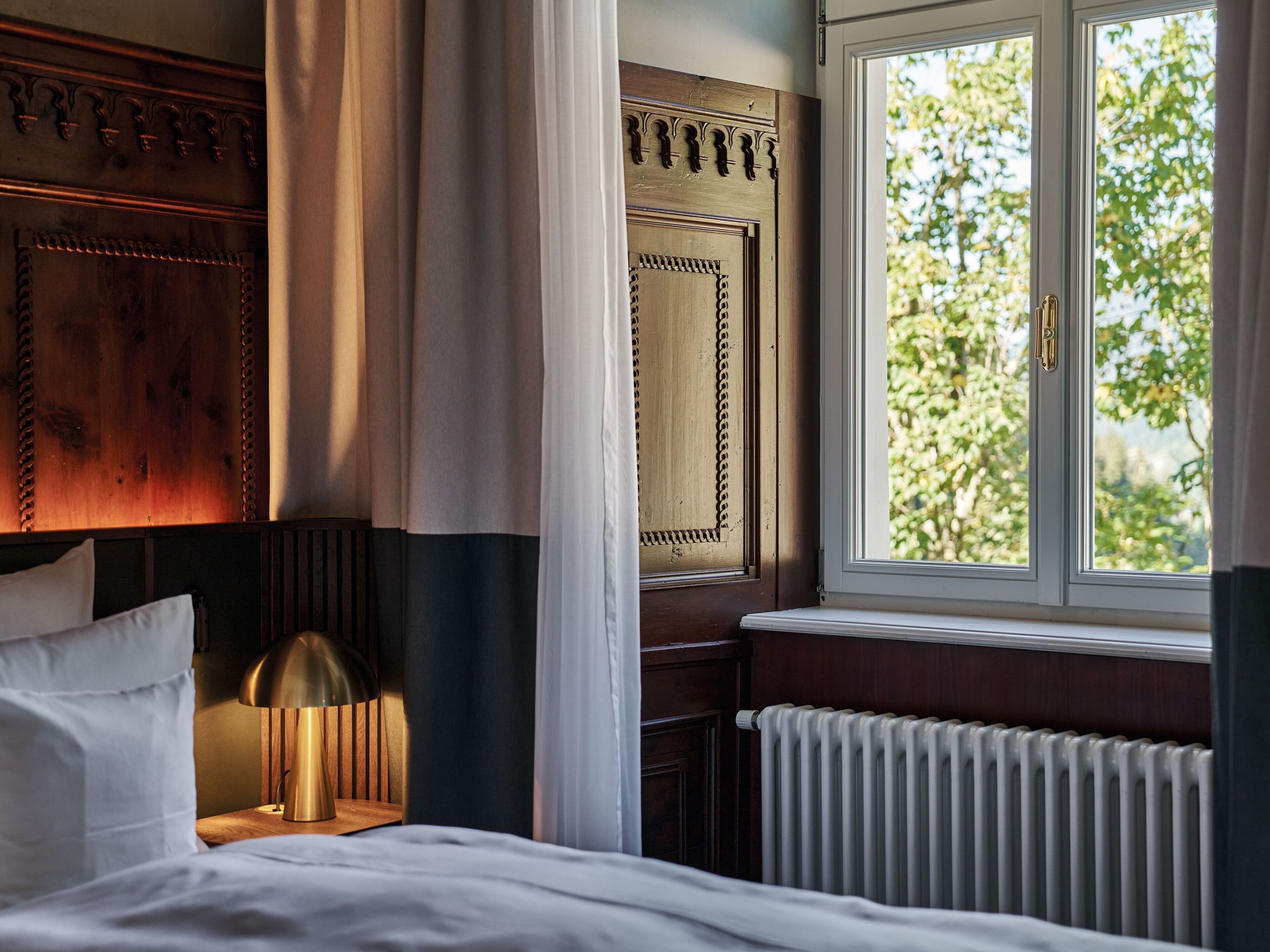 Una camera d'albergo con un letto, una lampada da comodino, una finestra vista alberi e un termosifone.
