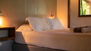 Ein Bett mit weißer Bettwäsche und zwei Kissen, ein Nachttisch aus Holz mit einer Lampe und ein Fenster mit Tageslicht.