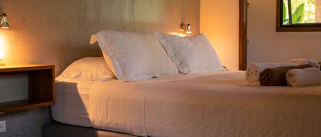 A bed with white linens and two pillows, a wooden nightstand with a lamp, and a window with daylight.