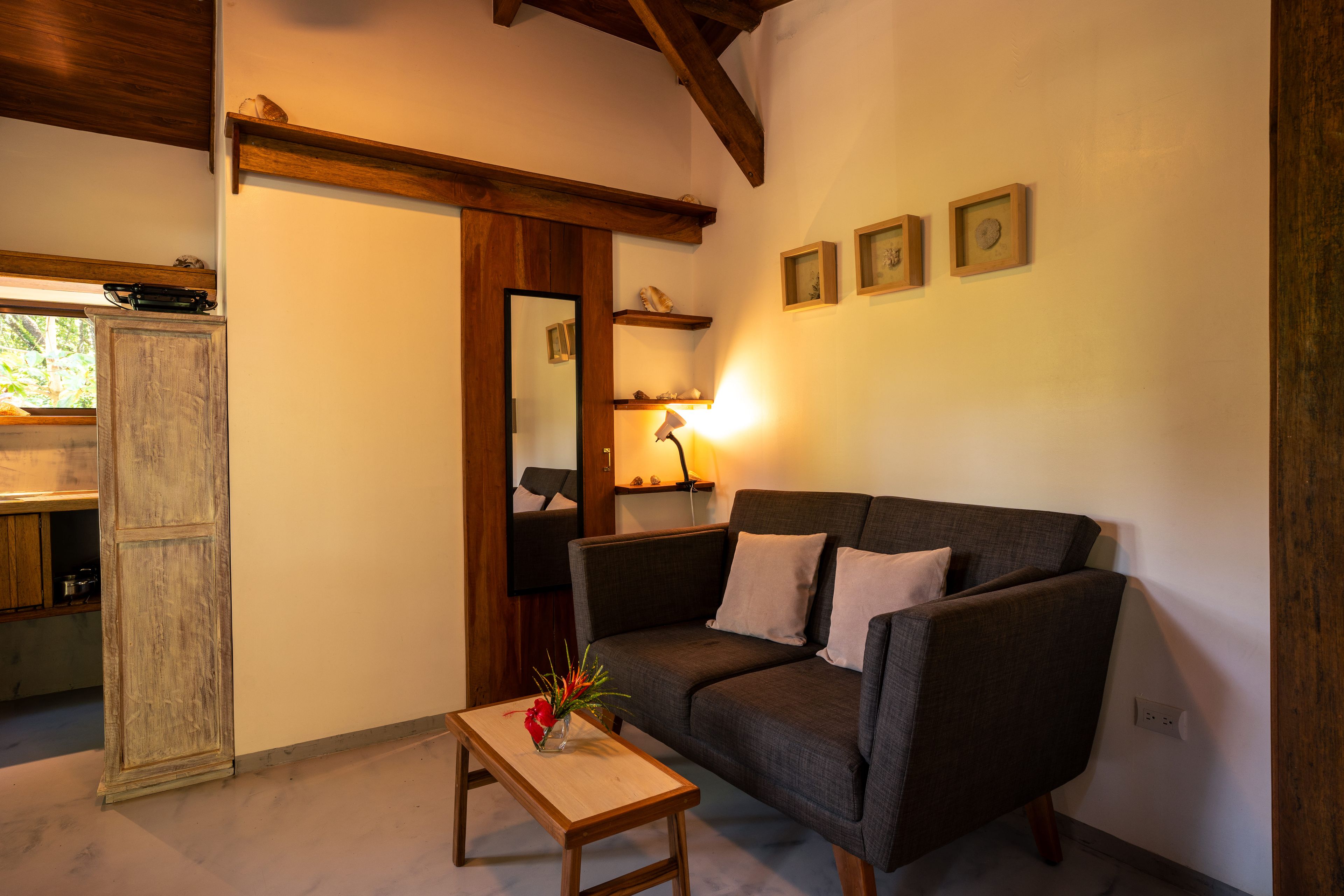 Una sala de estar acogedora con un sofá de color gris oscuro, una mesa de centro de madera con un jarrón, y una cocina visible a través de un puerta abierta.