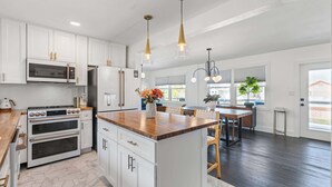 Una cocina con armarios blancos, electrodomésticos de acero inoxidable, una isla de madera, luces colgantes y un comedor con mesa y sillas.