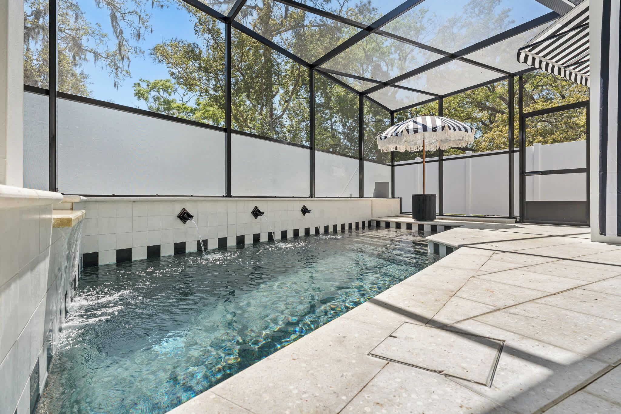 Ein Schwimmbecken mit klarem Wasser, umgeben von einer Glaskonstruktion, und eine Pergola mit einem gestreiften Sonnenschirm.