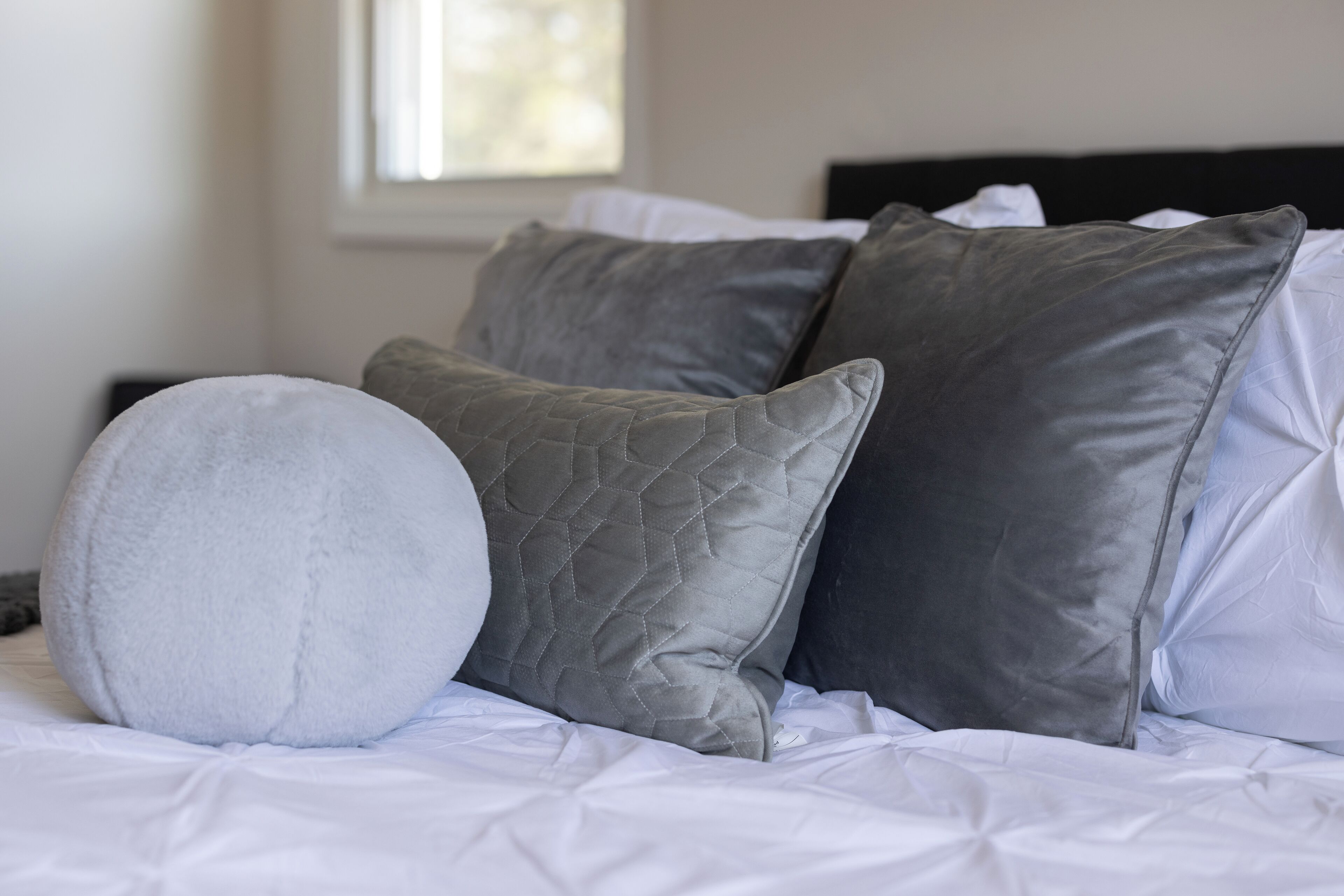 Un lit avec une literie blanche et plusieurs oreillers, dont un oreiller rond blanc et plusieurs oreillers gris.