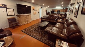 Ein Wohnzimmer mit einer braunen Ledersofa, einem Couchtisch und einem an der Wand befestigten Fernseher. Im Hintergrund ist eine Küche zu sehen.