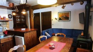 A cozy living room with a wooden dining table, blue sofa, and a kitchen area with a refrigerator and cabinets. - Ursula House - Your Mountain Holiday (Premana)